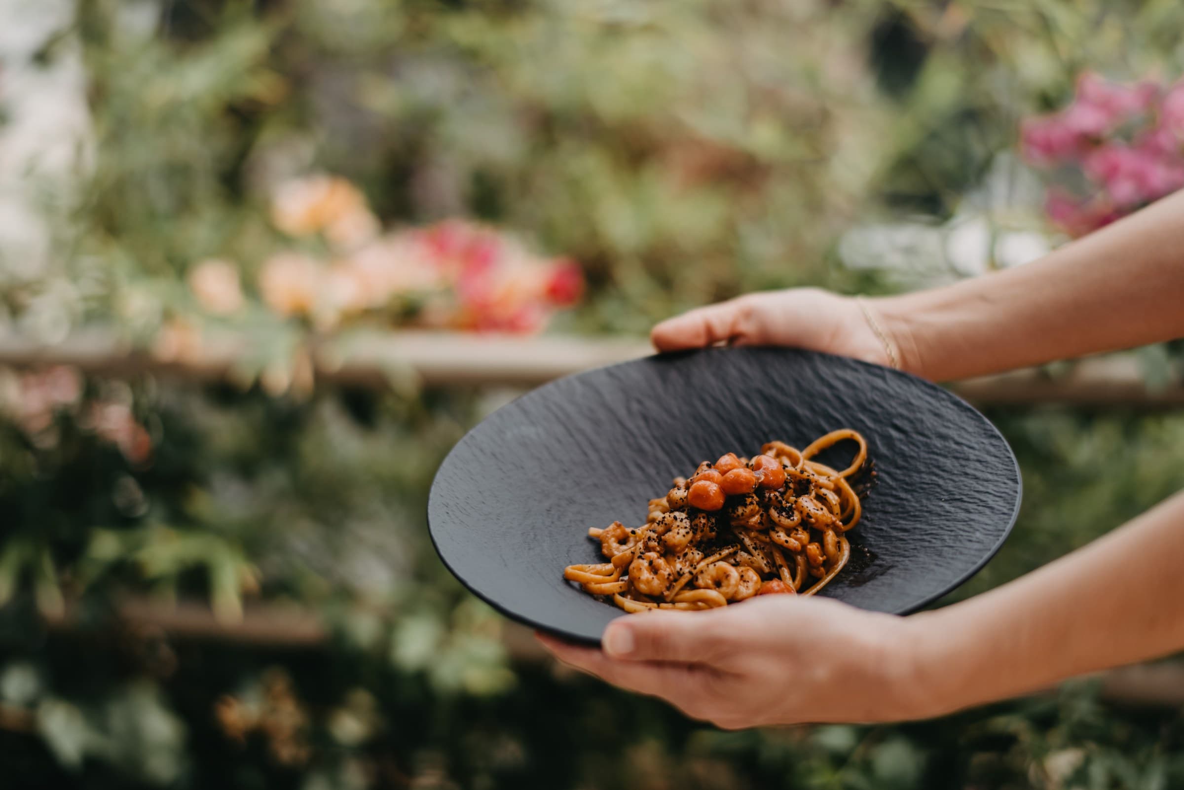 Patient hands carrying a plate of hand-cut pasta with Adriatic prawns through the garden of Zori Restaurant, Palmižana